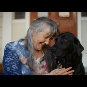 Jacqui is sitting and smiling with her guide dog in front of her house