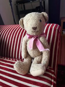 Image of a worn teddy bear with one eye missing and a pink bow around its neck