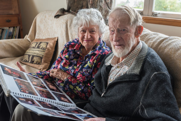 Hugh & Di on their couch with a photo album open on their laps smiling at the camera