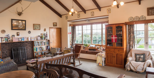 Hugh & Di's living room. There is a fireplace on the left, a couch in the middle of the room and a cabinet between 2 large windows that look out on the garden.