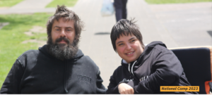 Bonnie (a young woman with short dark hair and using a wheelchair) beside a friend (a young man with a full, dark beard using a wheelchair) at National Camp 2023