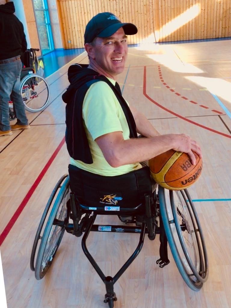 Ps Jack O. smiling at the camera in his wheelchair holding a basketball on a basketball court