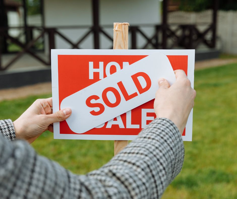 Two hands placing a "Sold" sticker on a house for sale sign.