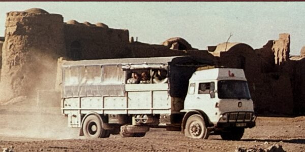 An old-style white cargo truck that has canvas sides in the rear with rectangular openings that serve as windows, which can be tied up for ventilation and to let passengers see out.