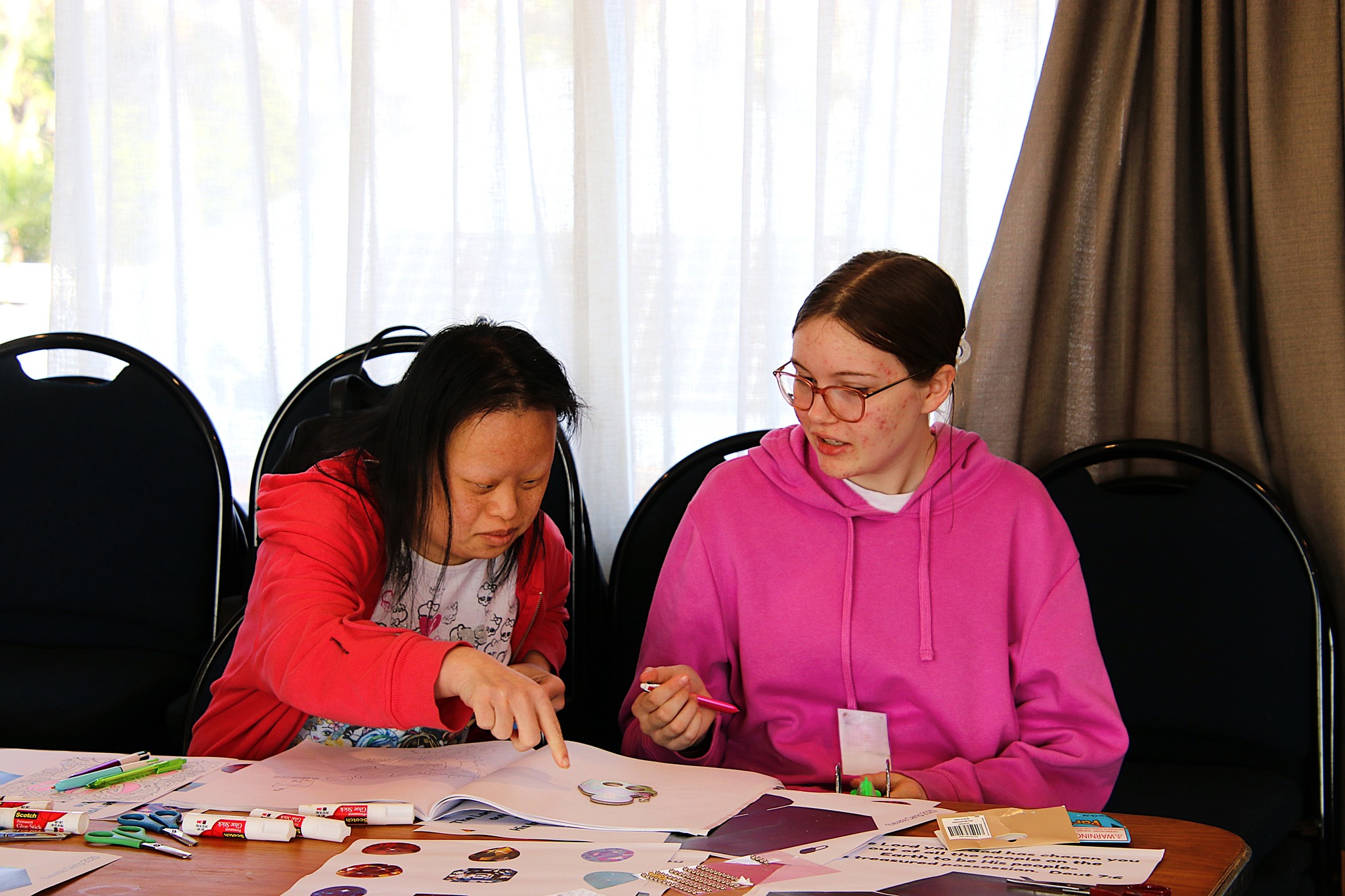 Photo of two young women doing crafts together inside, the table is covered in scrapbooking paper, glue and felt pens