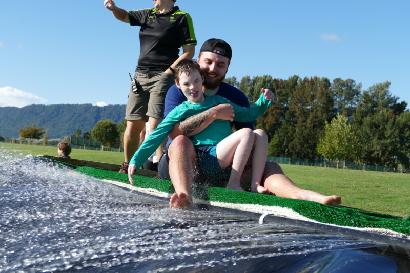 Photo of two young men, one camper and one volunteer, both sitting at the top of the waterslide ready to go down