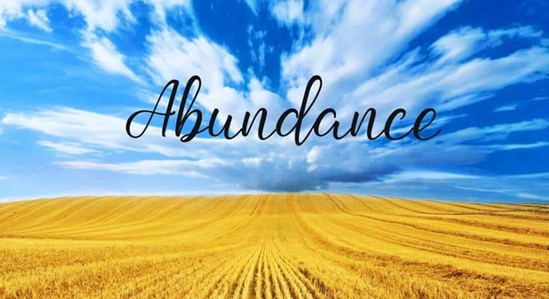 A field of hay slopes into the distance with bright blue sky above. The word abundance is scrawled across the sky.