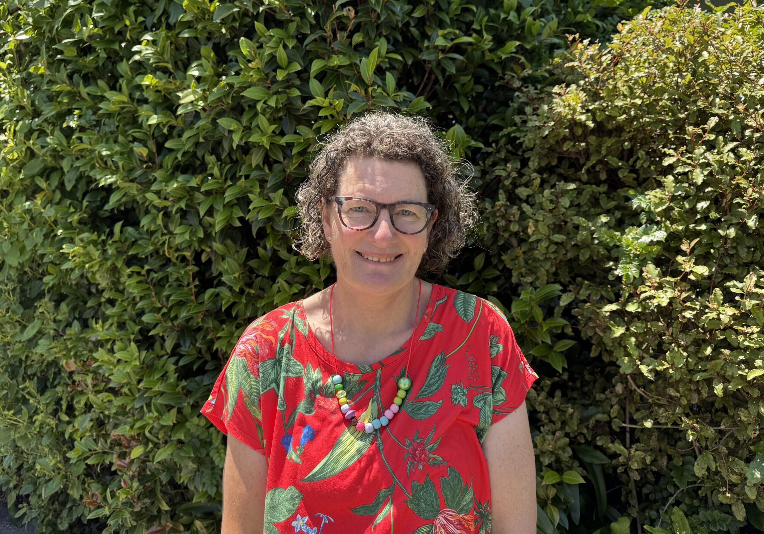 Alison has brown curly hair, wears glasses, a red floral shirt and a beautiful, beaded necklace.