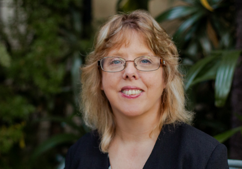 Christine has blonde curly hair and wears glasses. She's wearing a floral-patterned shirt.