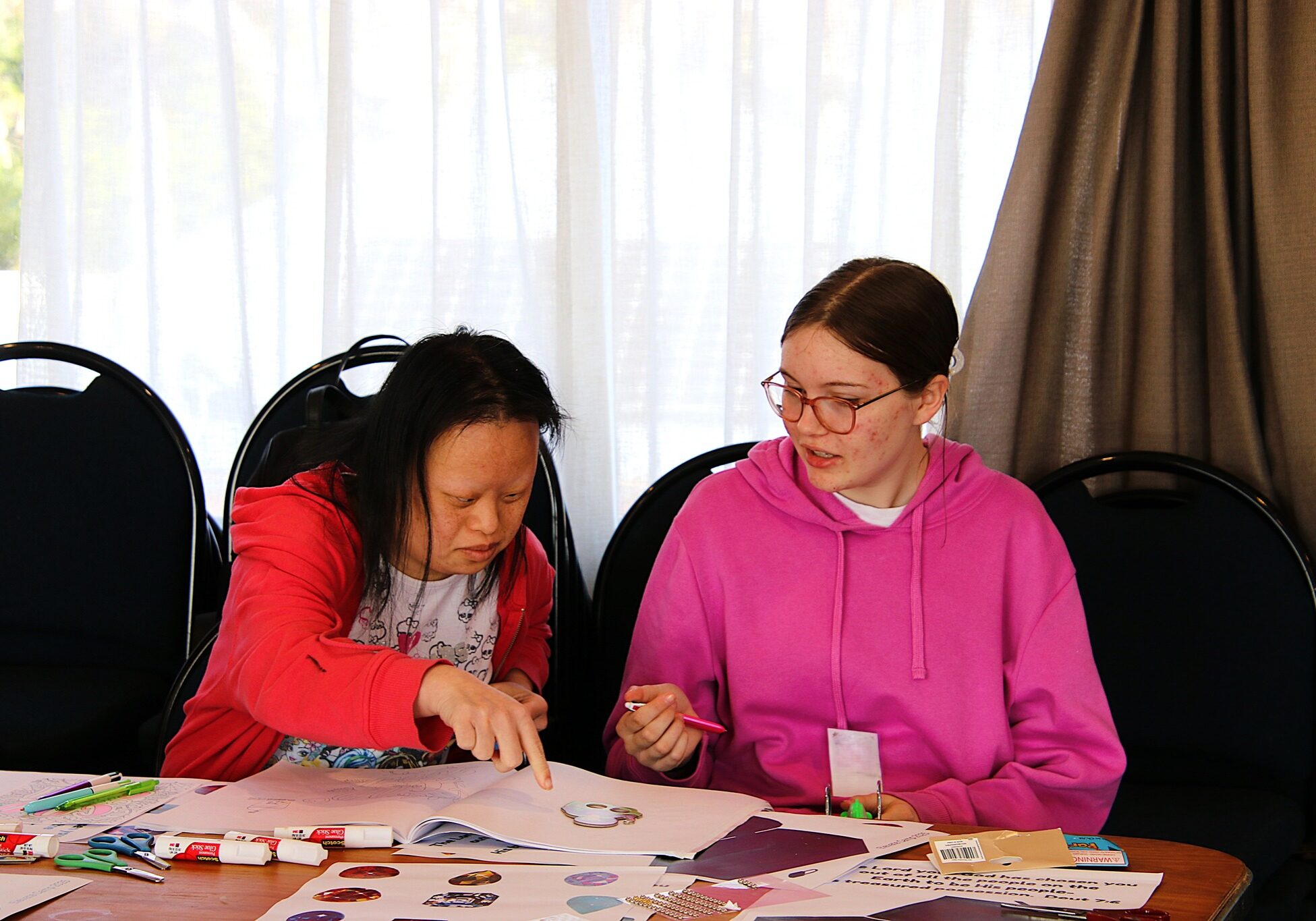 Photo of two young women doing crafts together inside, the table is covered in scrapbooking paper, glue and felt pens