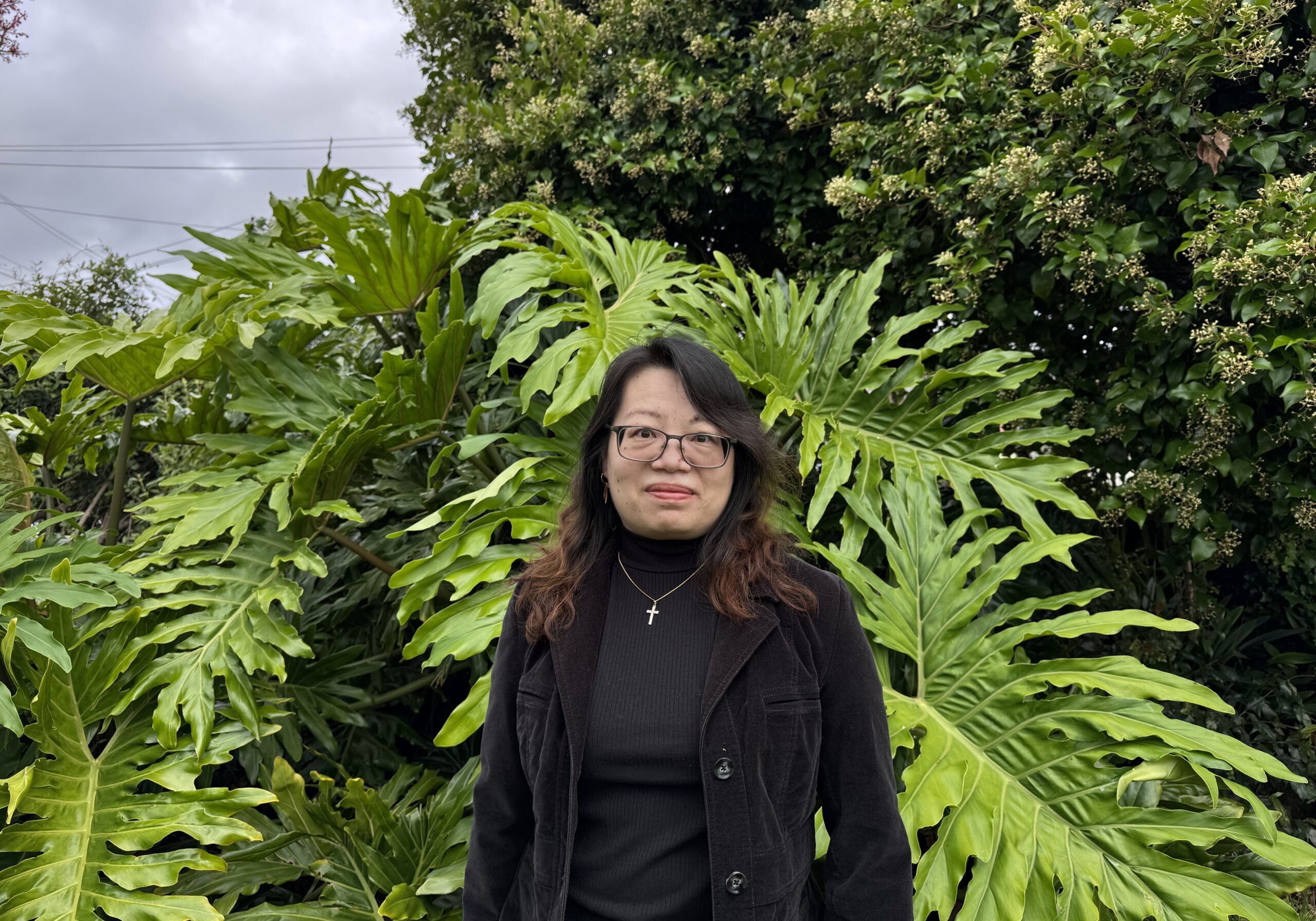 Jacqui has long black hair and wears a black shirt and jacket, with a silver cross necklace.