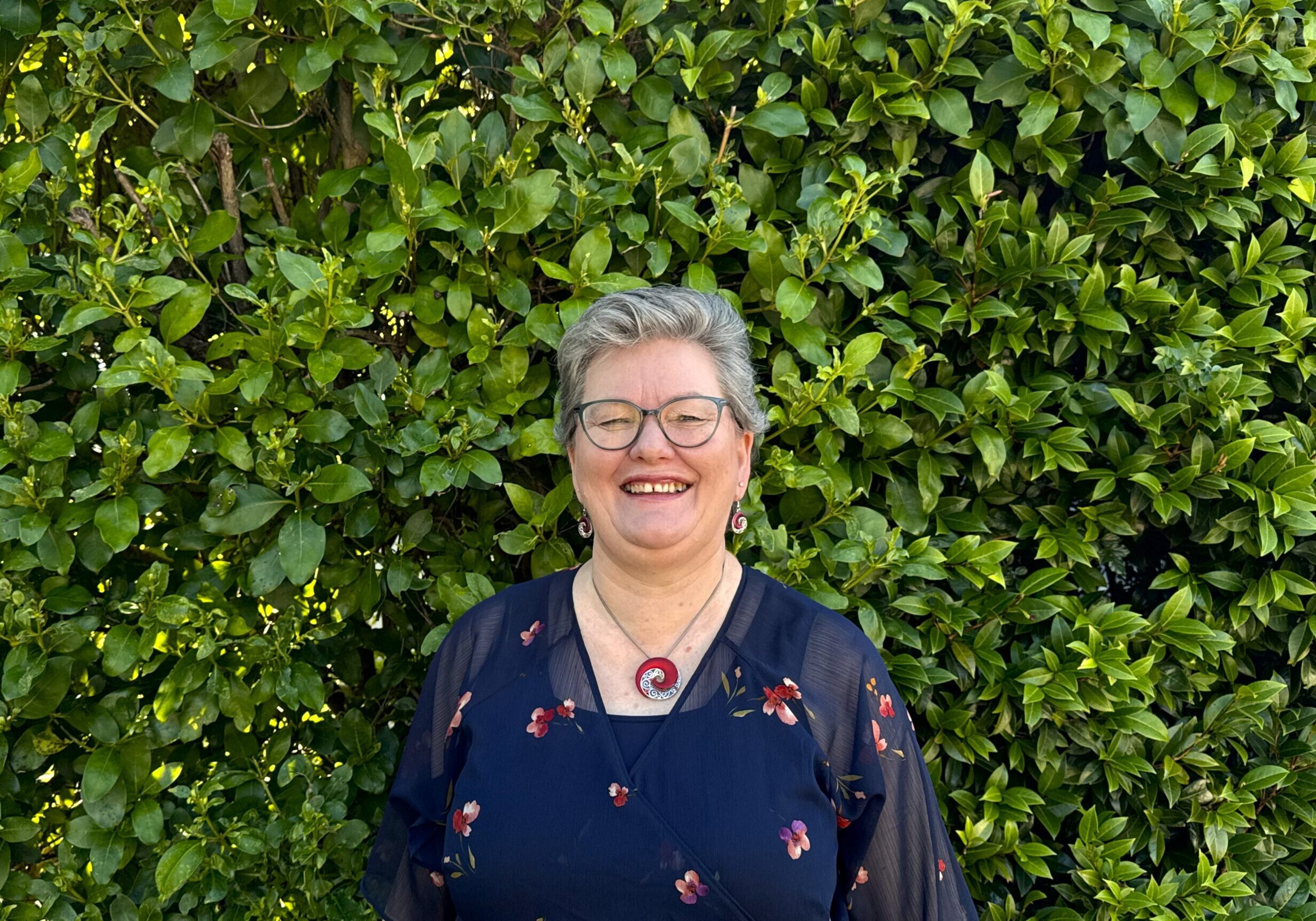 Kathie has short hair, wears glasses, and has a lovely red necklace that is shaped like a koru. She's wearing a flowing blue dress.