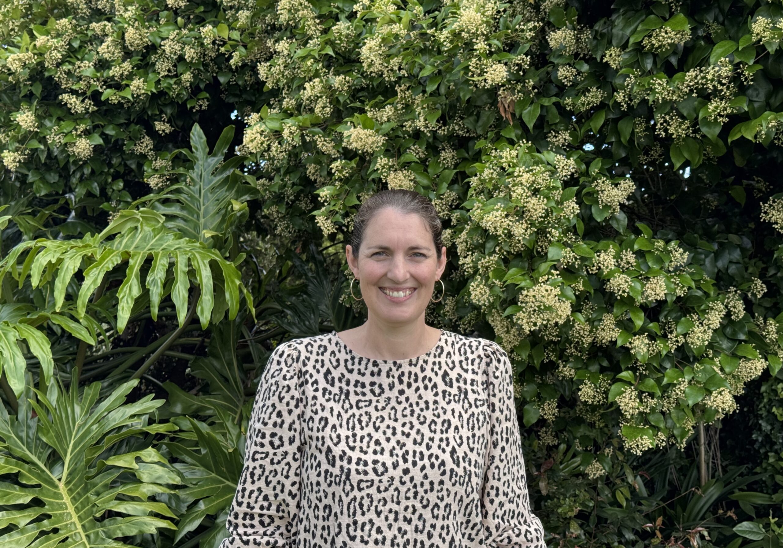 Kirsty Armitage, Director, wears a leopard print shirt and has brunette hair. Kirsty stands outside, in front of the centre's garden.