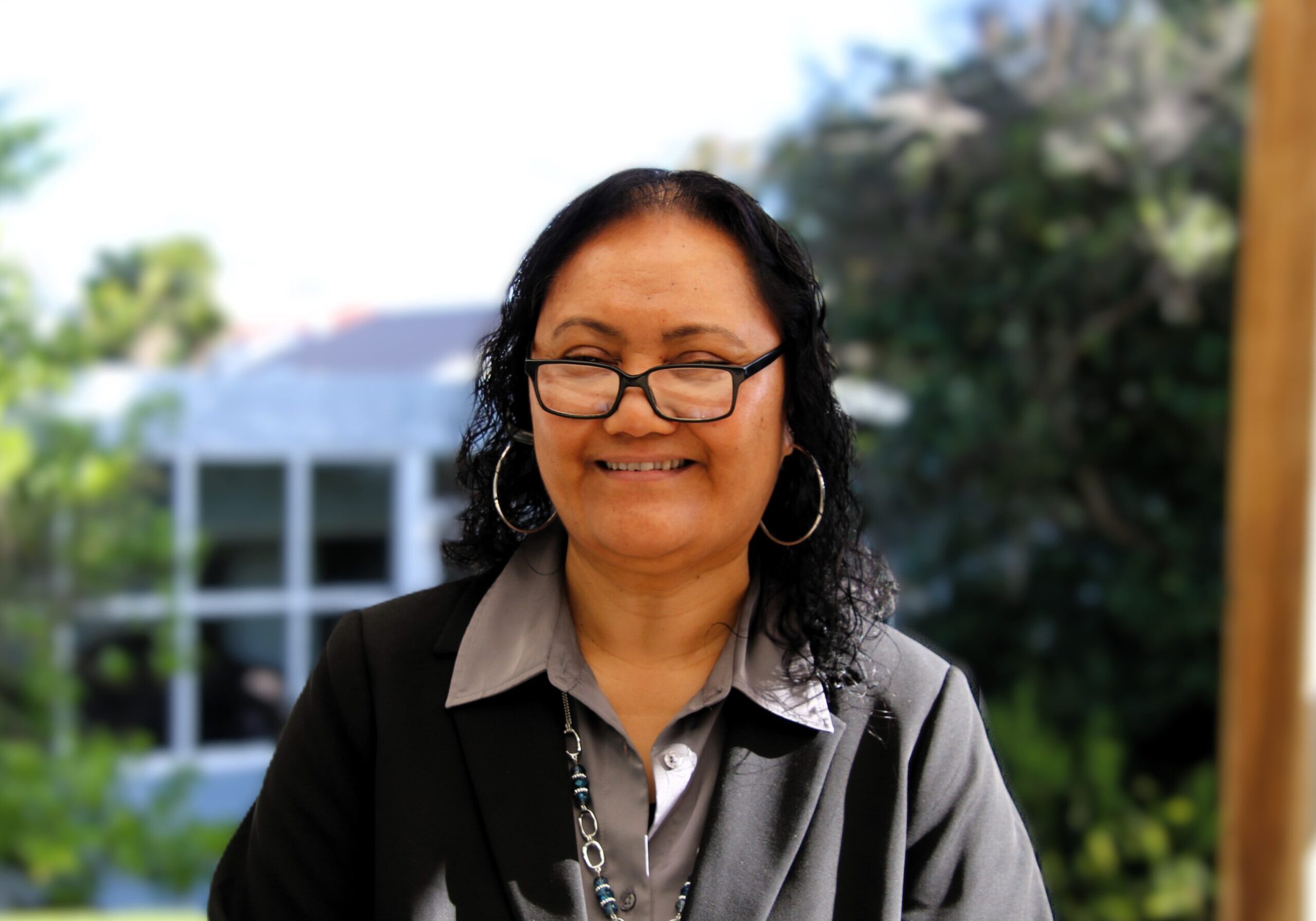 Teremoana has short black curly hair, wears glasses, hoop earrings, and a black button-up cardigan.